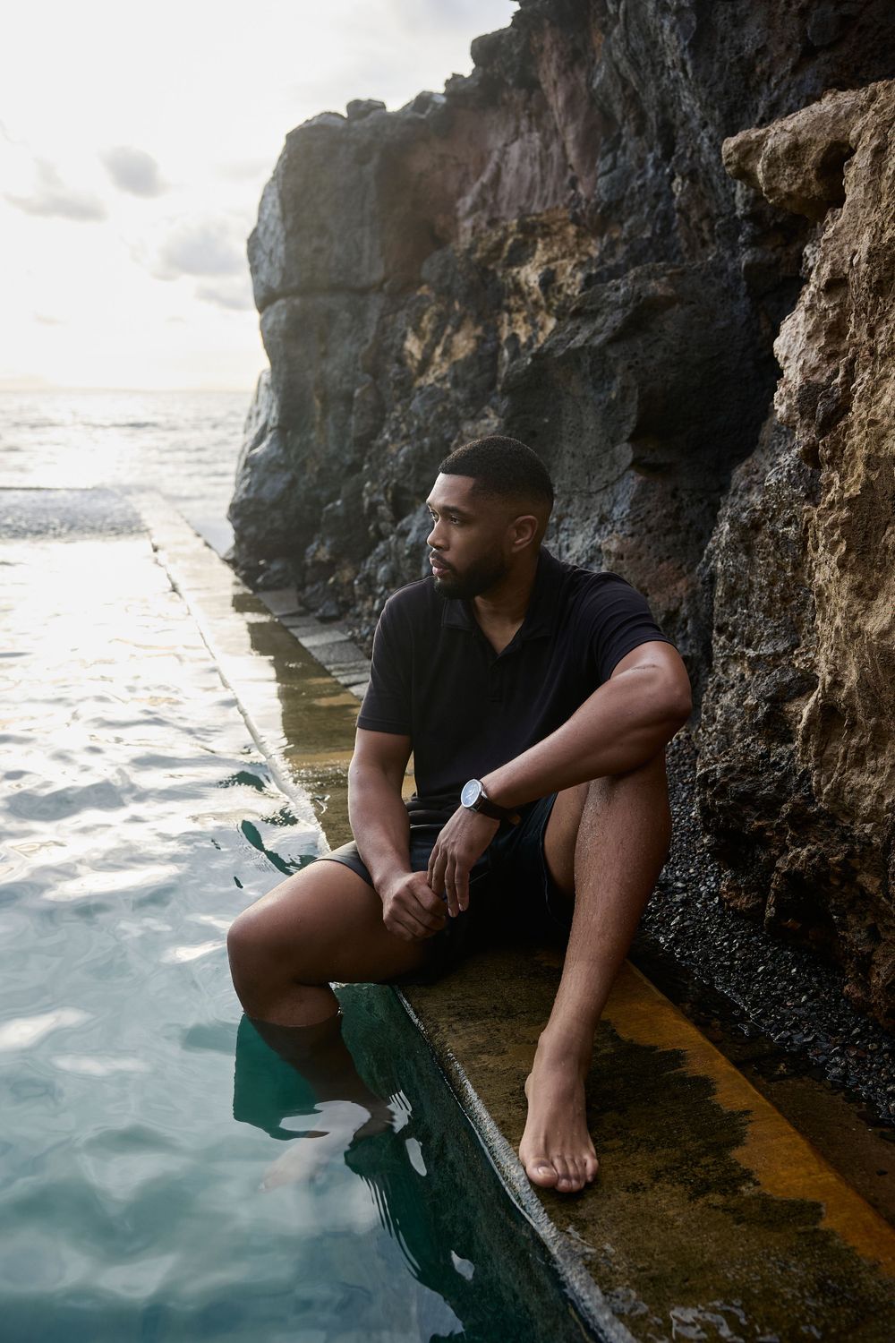 Man sitting by ocean pool at sunset, leaning against rocky cliff in black polo and shorts, peaceful and reflective vibe.