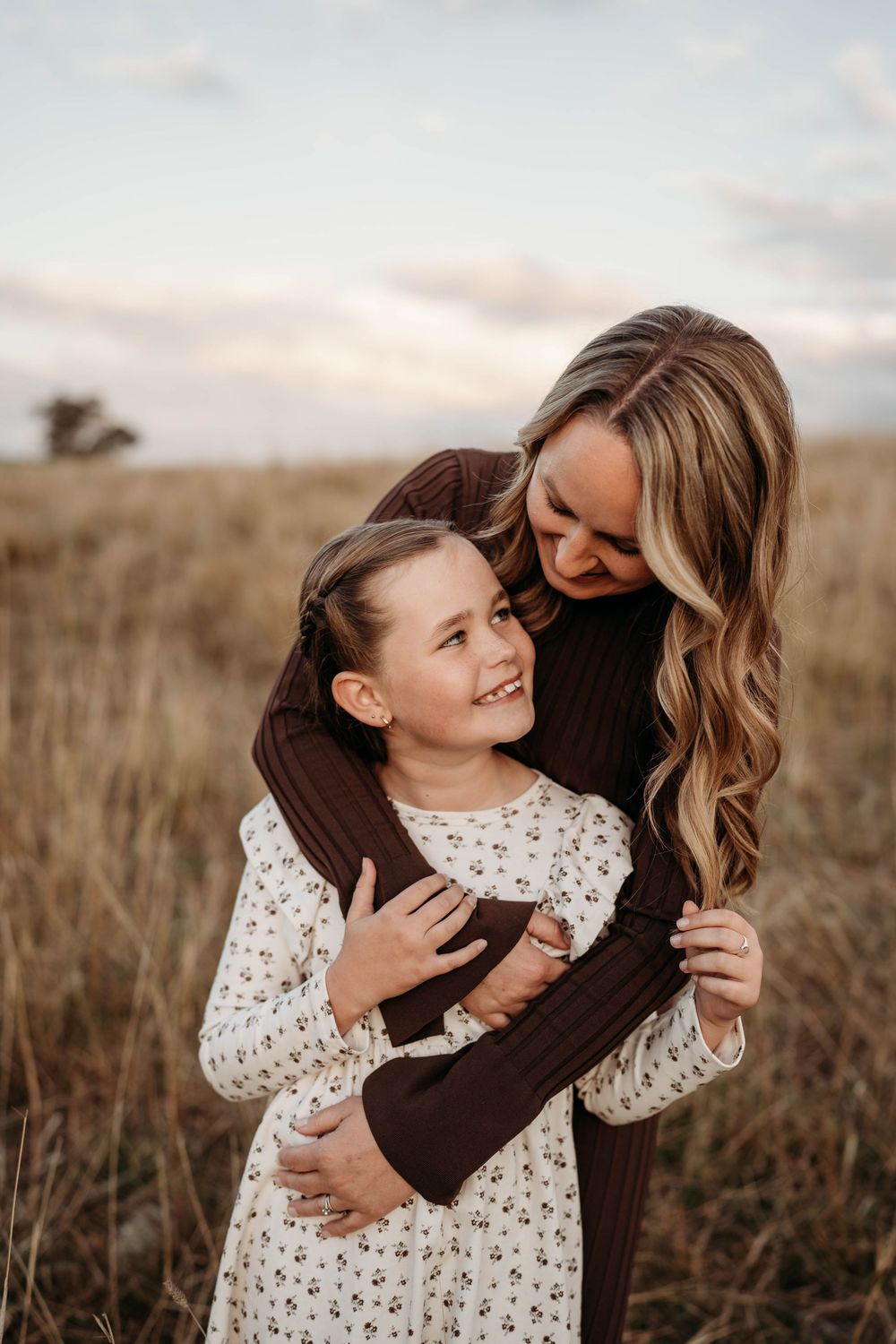 Mother in brown sweater hugging smiling daughter in floral dress in autumn golden field.