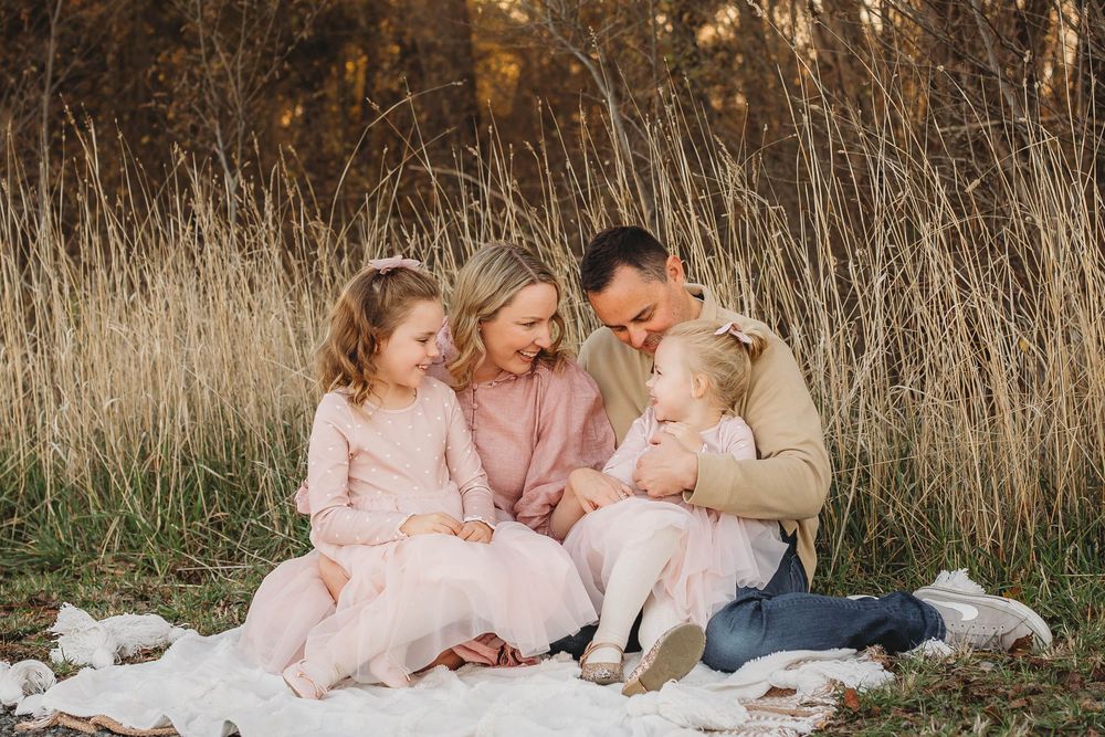Family of four with two girls in pink dresses sitting on blanket in tall grass field.
