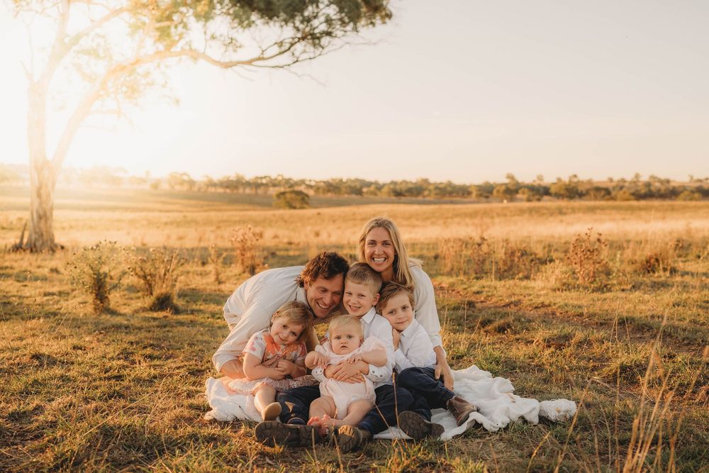 Happy family of five sitting in golden field at sunset, parents with three young children smiling.