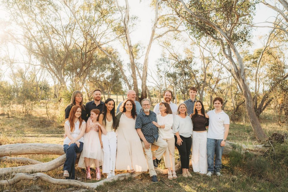 Extended family of thirteen smiling together in a sun-drenched eucalyptus woodland setting.