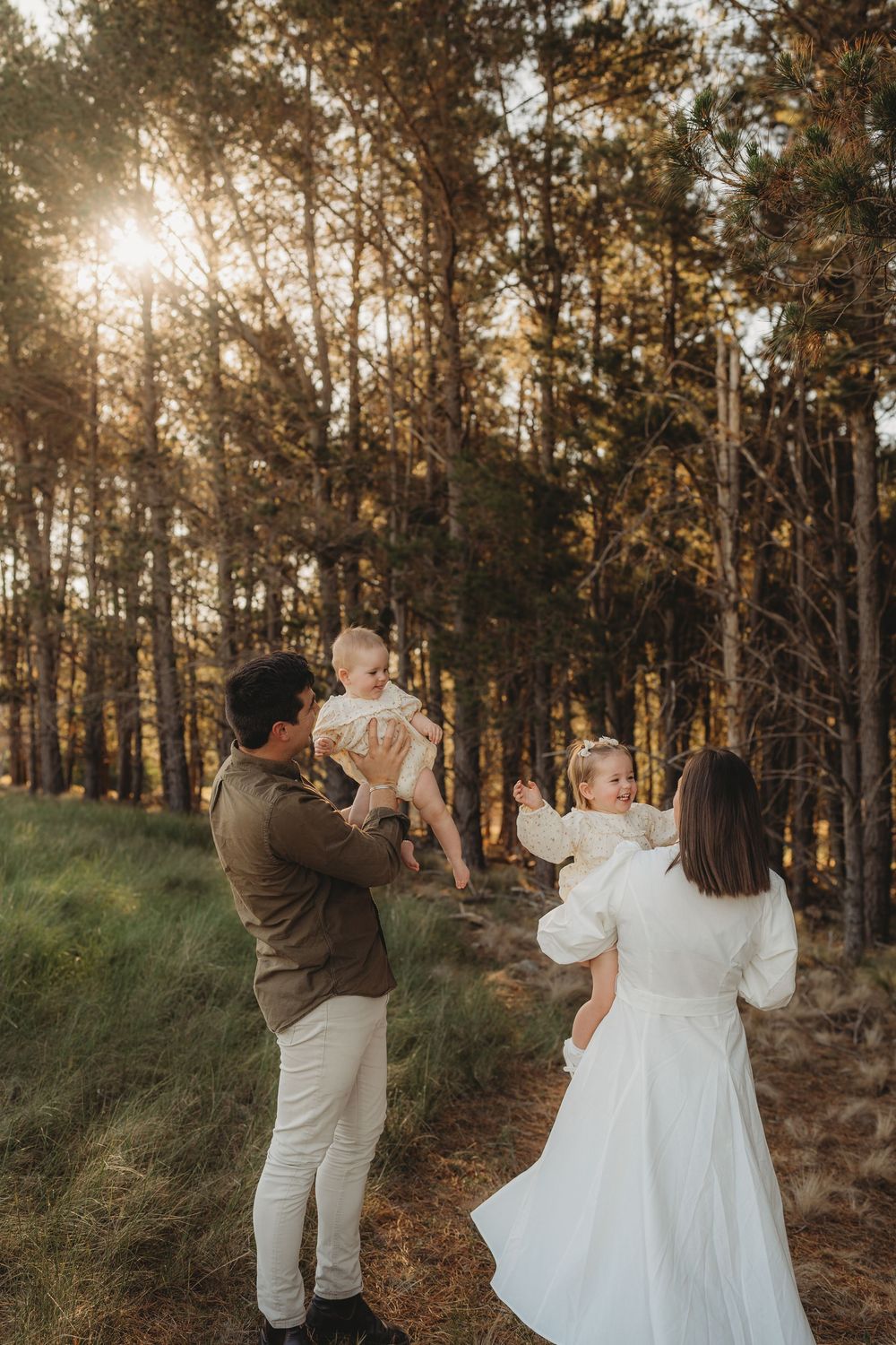 Parents lifting twin babies in sunlit pine forest, joyful family outdoor portrait at golden hour.