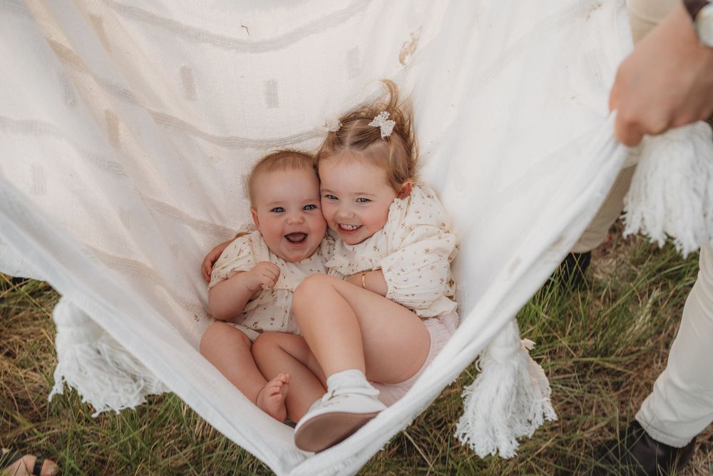 Two happy baby sisters cuddling together in a white fabric hammock on green grass.