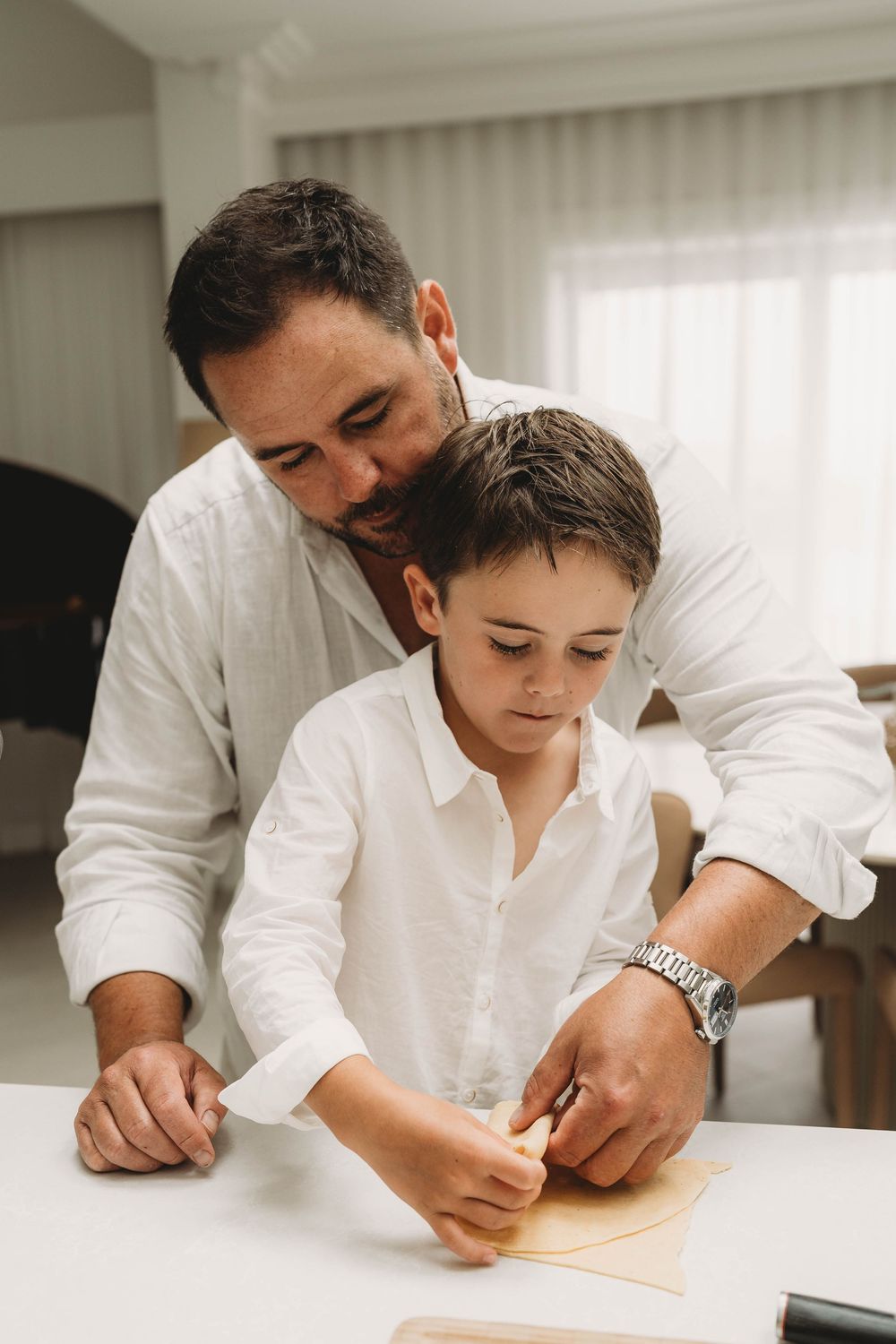 Father and young son working together on a craft project at a white table indoors.