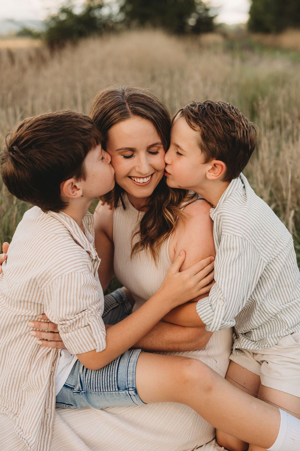Two boys kissing their smiling mother on the cheeks while sitting in a grassy field.