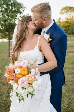 Romantic wedding couple embraces in outdoor setting with vibrant orange and pink floral bouquet at sunset.