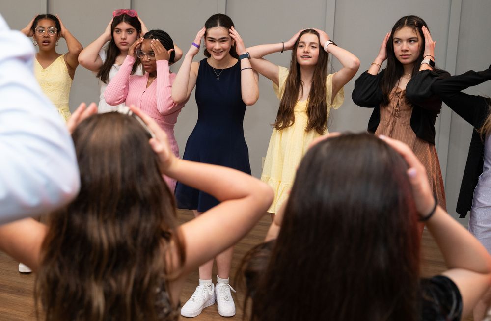 Young girls rehearsing a dance routine in a studio, following an instructor's movements with hands on heads.