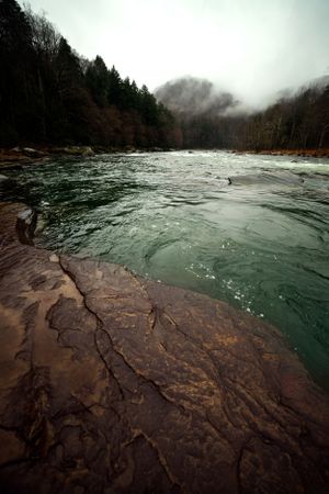 Dramatic view of a rushing river with misty mountains and forest in the background.