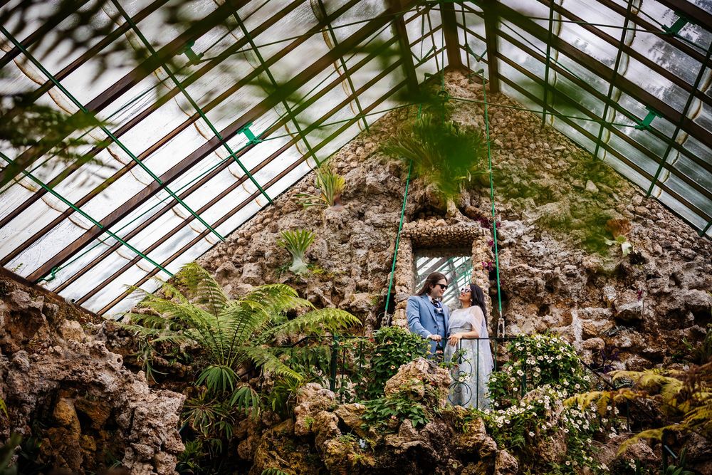Two people stand together on a stone path inside a sunlit greenhouse filled with rocks and tropical plants.