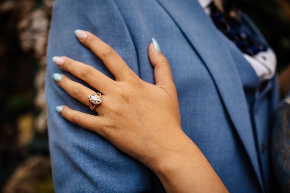 A well-manicured hand wearing an engagement ring rests against a navy blue suit jacket captured during an engagement shoot by Lynette