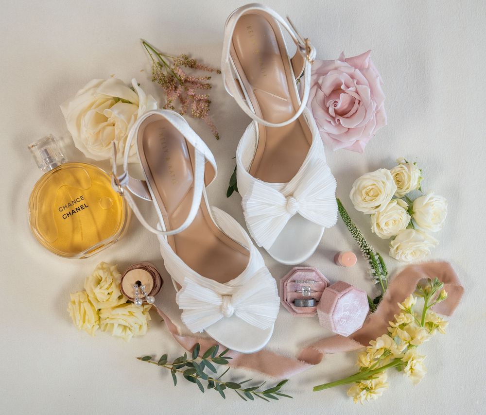 White bridal heels with bow detail surrounded by roses, Chanel perfume, and pink flowers on a white background.