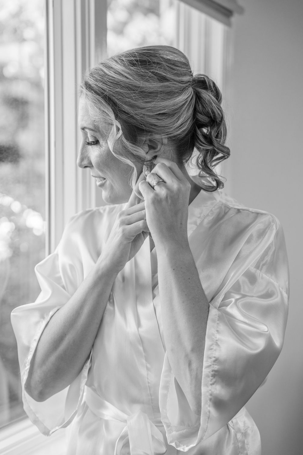 Bride in silk robe putting on earrings by window in black and white wedding preparation photo.