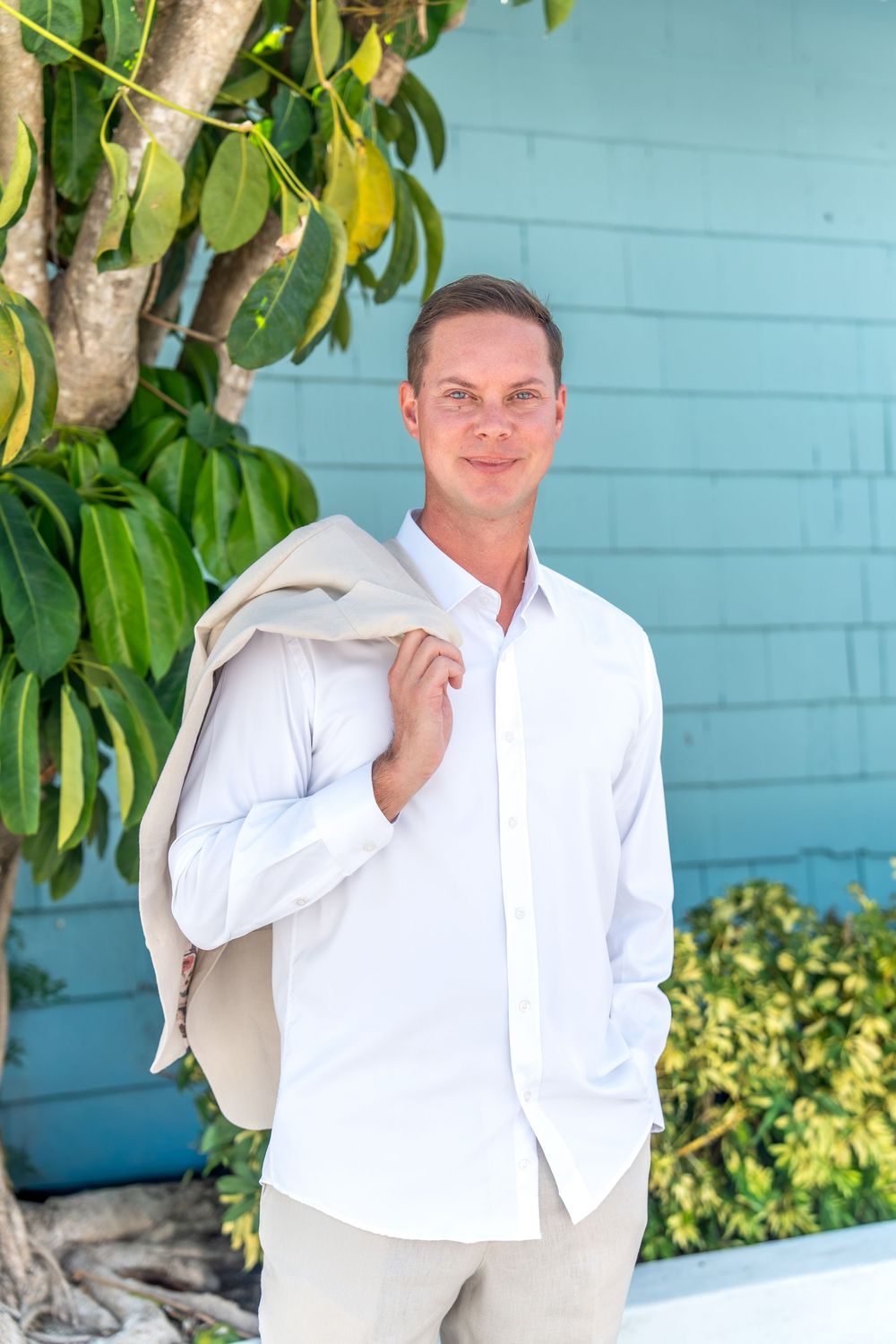 Man in white shirt holding beige blazer over shoulder, smiling outdoors near tropical tree and teal wall.