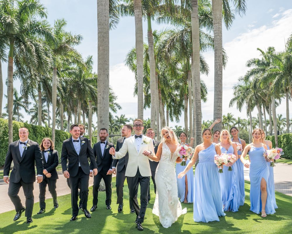 Wedding party walks together outdoors among tall palm trees, bridesmaids in light blue gowns and groomsmen in black tuxedos.
