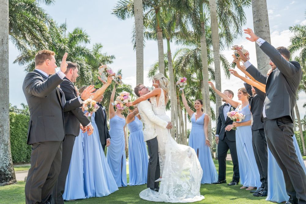 Wedding party celebrating couple kissing outdoors, bridesmaids in blue gowns and groomsmen in gray suits, tropical palm trees background.