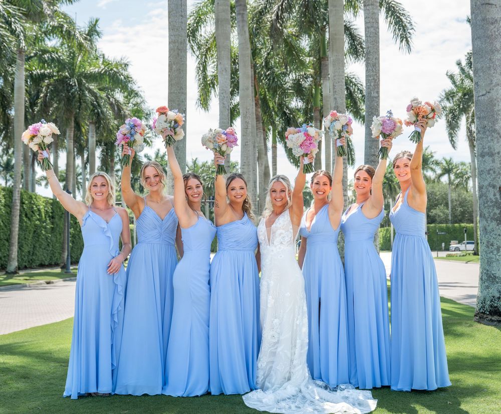 Bridesmaids in matching light blue gowns raise bouquets with bride outdoors surrounded by tropical palm trees.