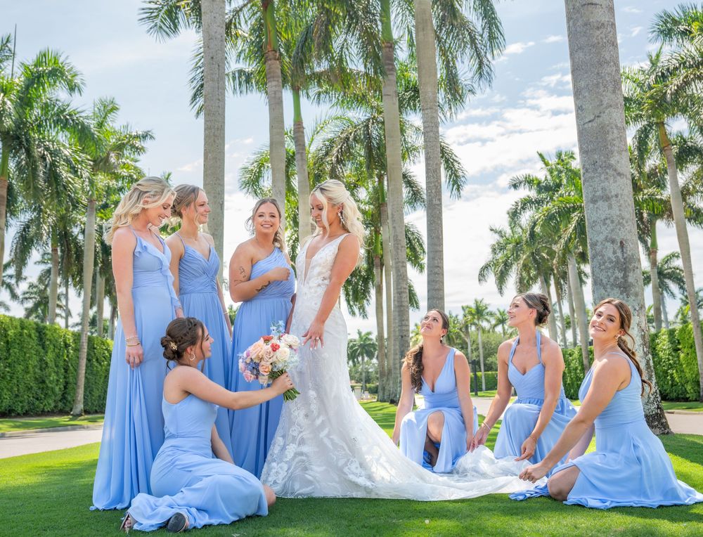 Bride in white gown surrounded by bridesmaids in blue dresses among tropical palm trees outdoors.