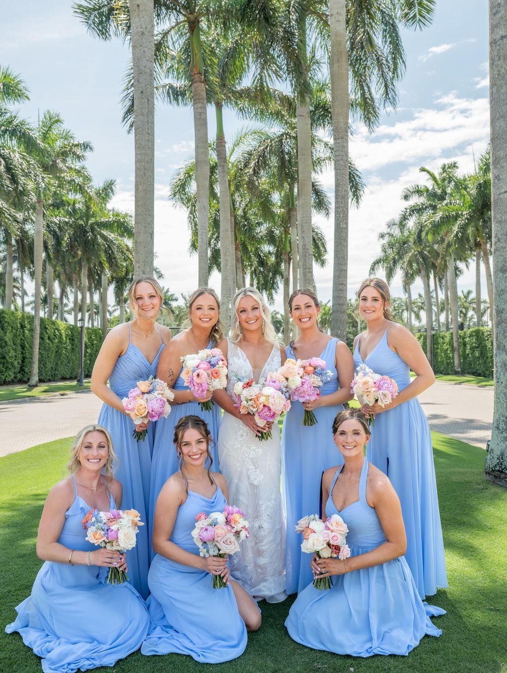 Bride in white gown poses with seven bridesmaids in light blue dresses holding pink bouquets among tall palm trees.