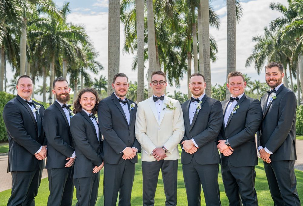 Groom in white tuxedo jacket poses with seven groomsmen in dark suits and bow ties outdoors with palm trees.