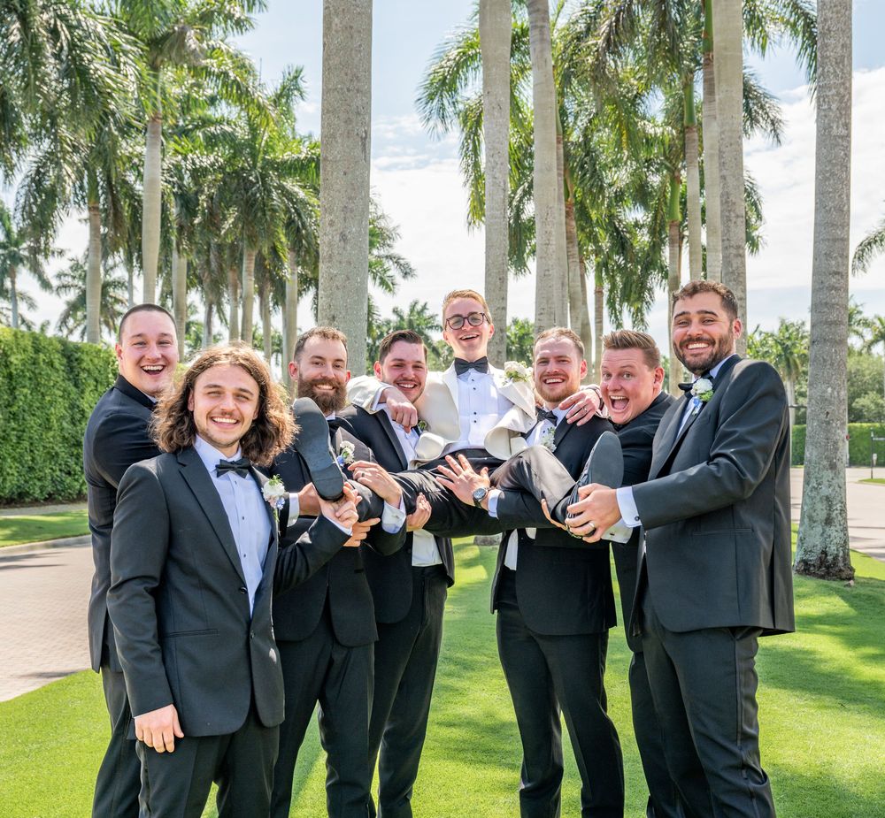Groomsmen in black tuxedos playfully lift groom outdoors surrounded by tropical palm trees.