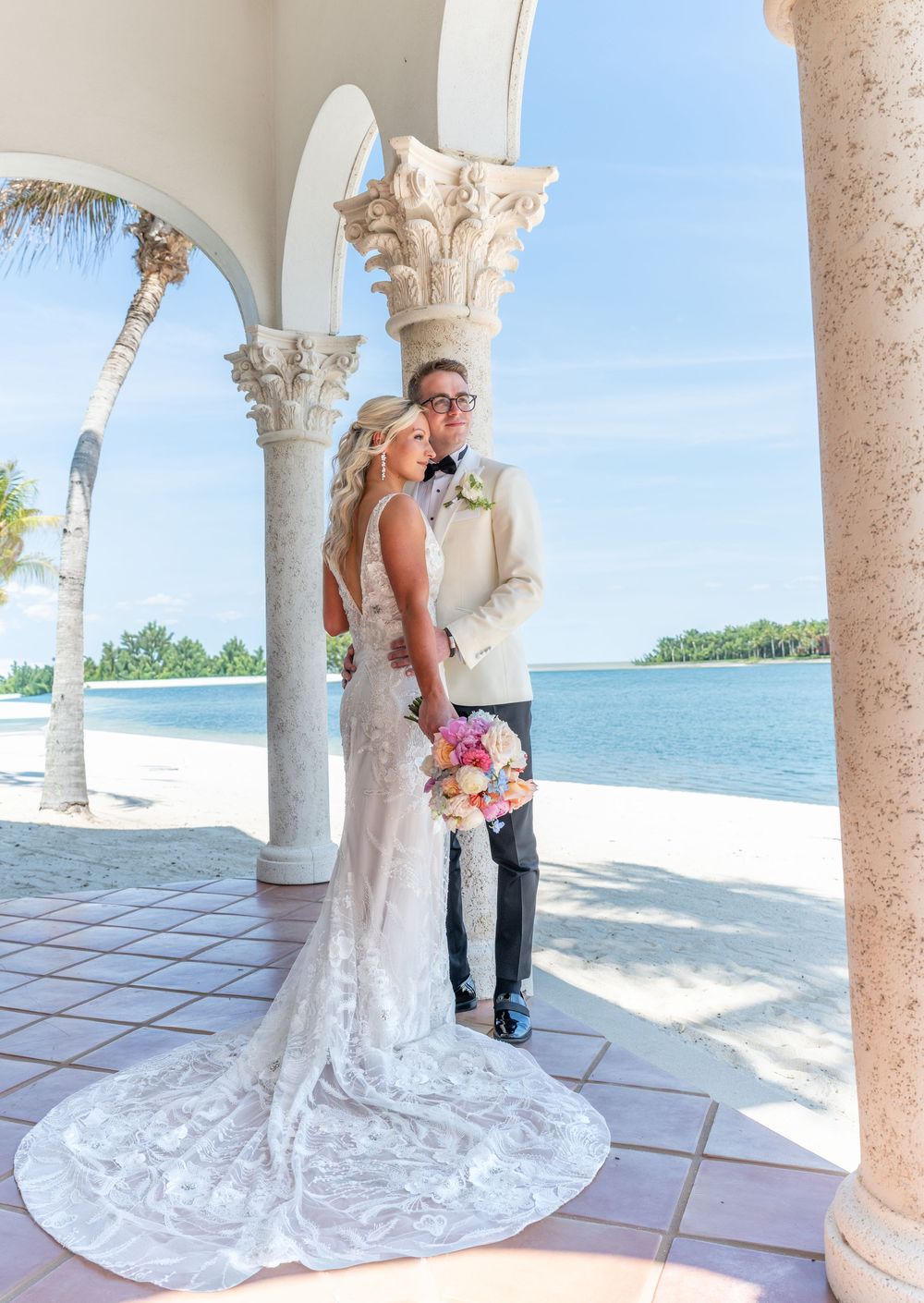 Bride and groom pose under elegant arched columns with a stunning beachside ocean view on their wedding day.