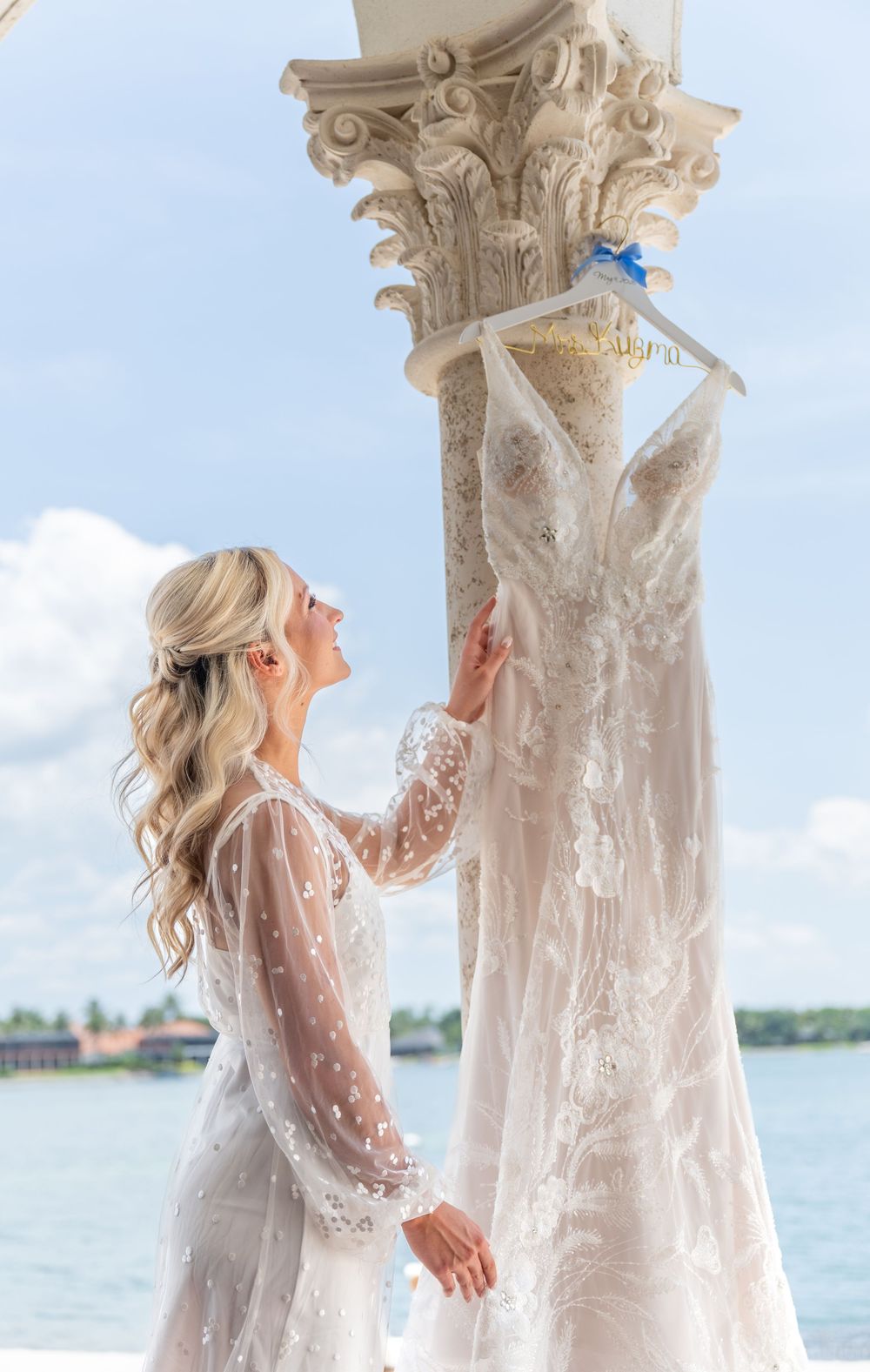 Blonde bride in lace robe admires her beaded wedding gown hanging on a decorative column by the waterfront.