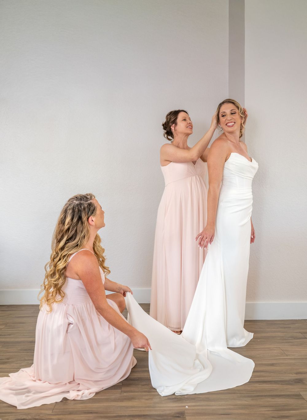 Two bridesmaids in blush pink gowns help a smiling bride in a white wedding dress prepare before her ceremony.
