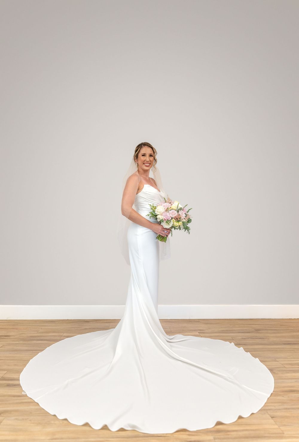 Bride in elegant white satin mermaid wedding gown with long train holds pink bouquet, posing in bright studio.