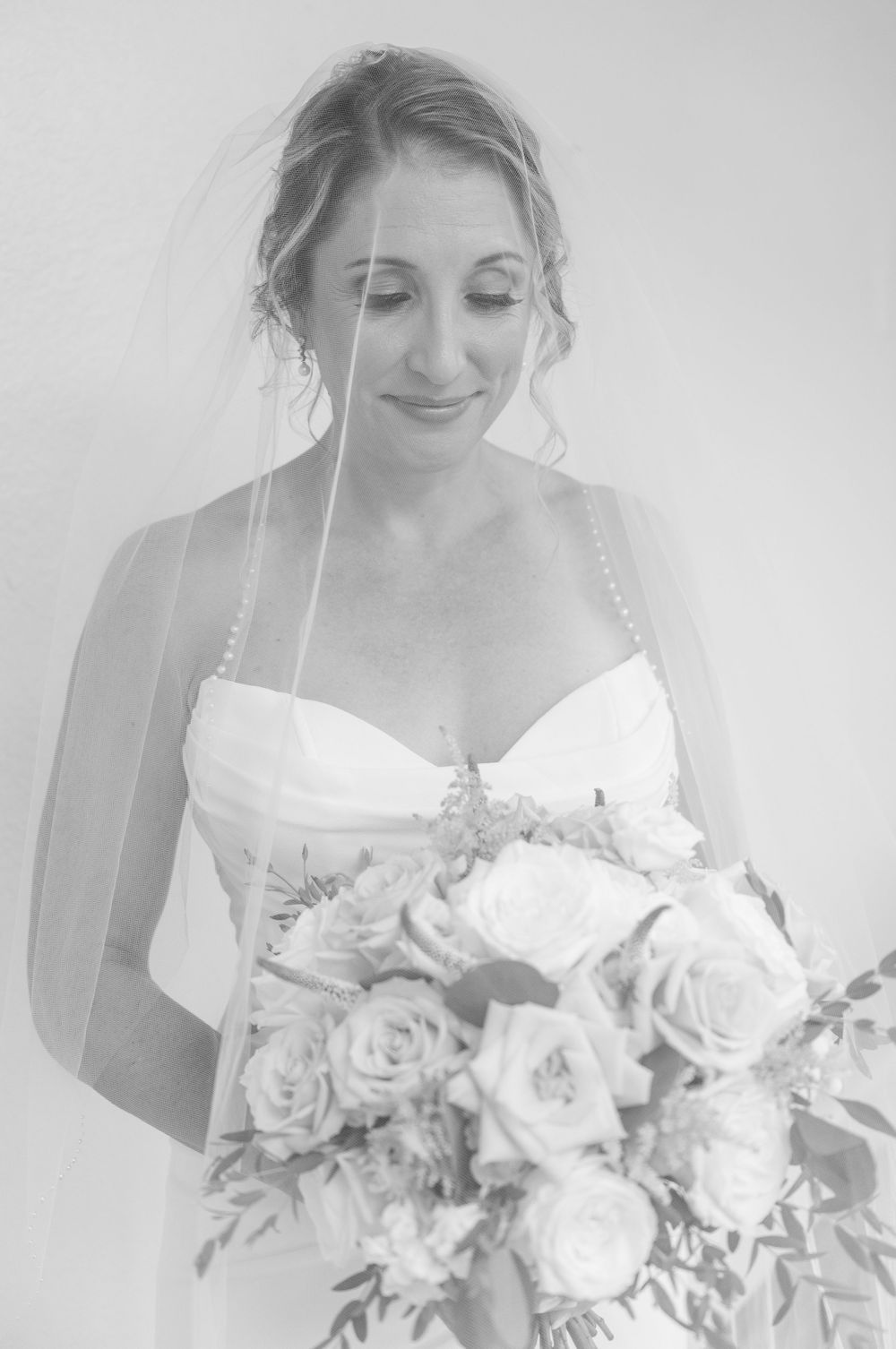 Black and white portrait of a smiling bride in a strapless gown holding a rose bouquet with a veil.