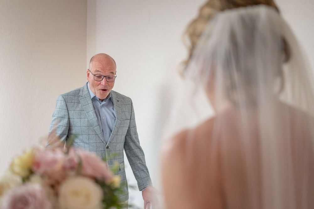 A bald man in a gray suit reacts with awe seeing a bride in a white gown and veil during a wedding first look.