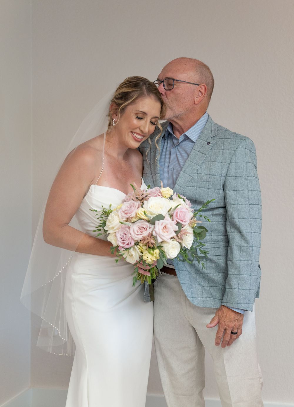 Bride in white gown holding pink rose bouquet receives a kiss on cheek from older man in light blue plaid blazer.
