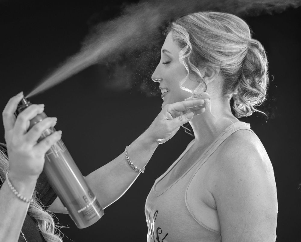 Black and white photo of a bride getting hairspray applied to her updo hairstyle during wedding preparations.