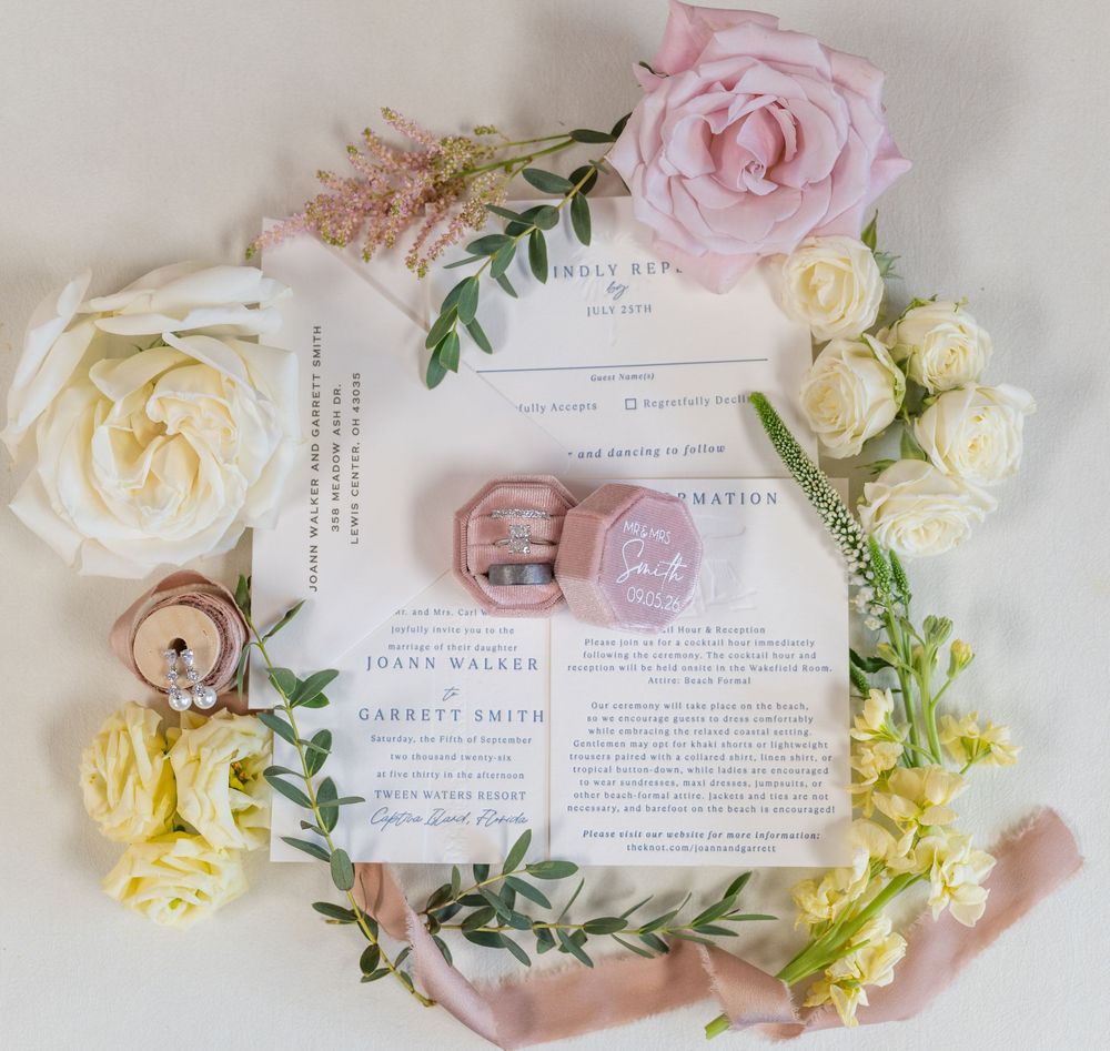 Flat lay of wedding invitations surrounded by pink and cream roses with a pink ring box and ribbon.