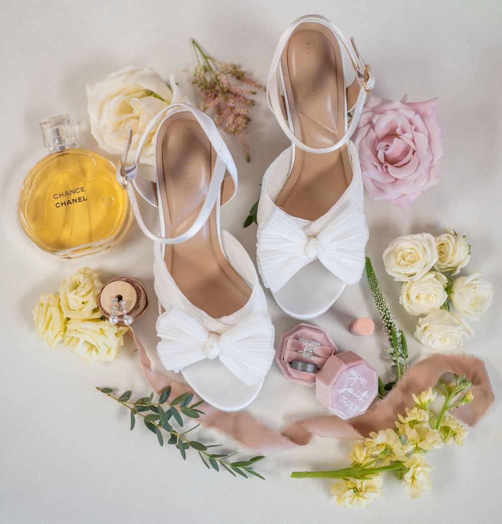 White bridal heels with bow detail surrounded by roses, Chanel perfume, and pink accessories on a white background.
