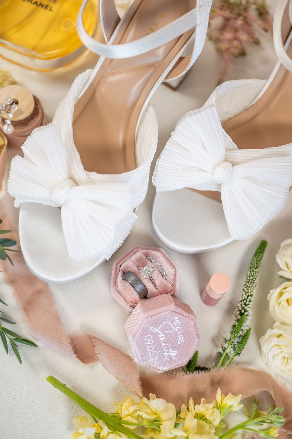 White bow bridal heels displayed with pink ring box, pearl earrings, Chanel perfume, and floral accents on a wedding flat lay.