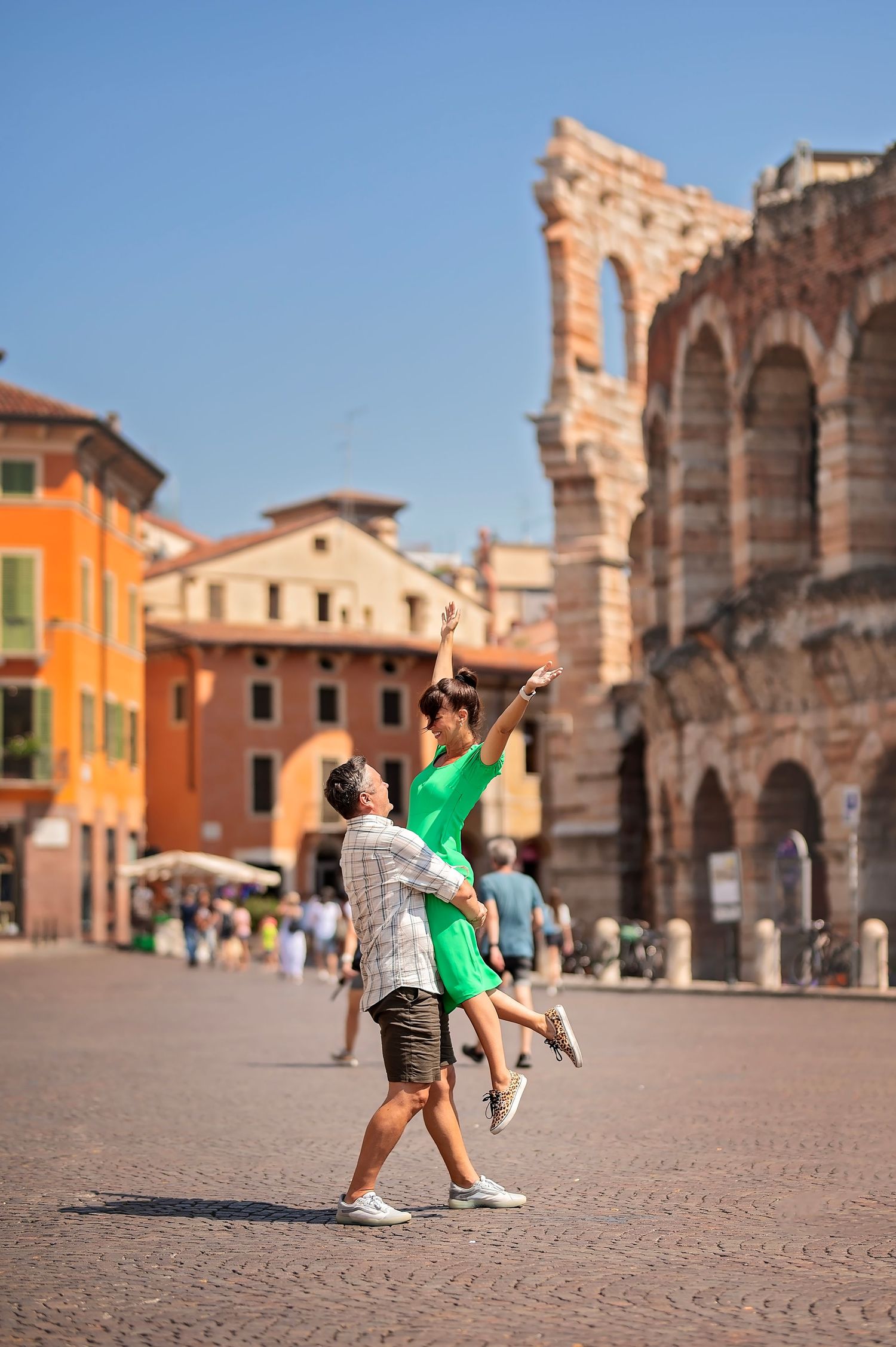 Joyful couple performing lift pose in front of historic Italian amphitheater on sunny day.
