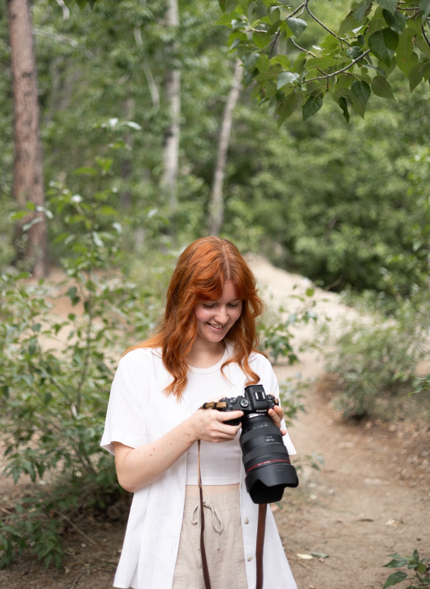 Redhead woman reviewing photos on DSLR camera on wooded path.