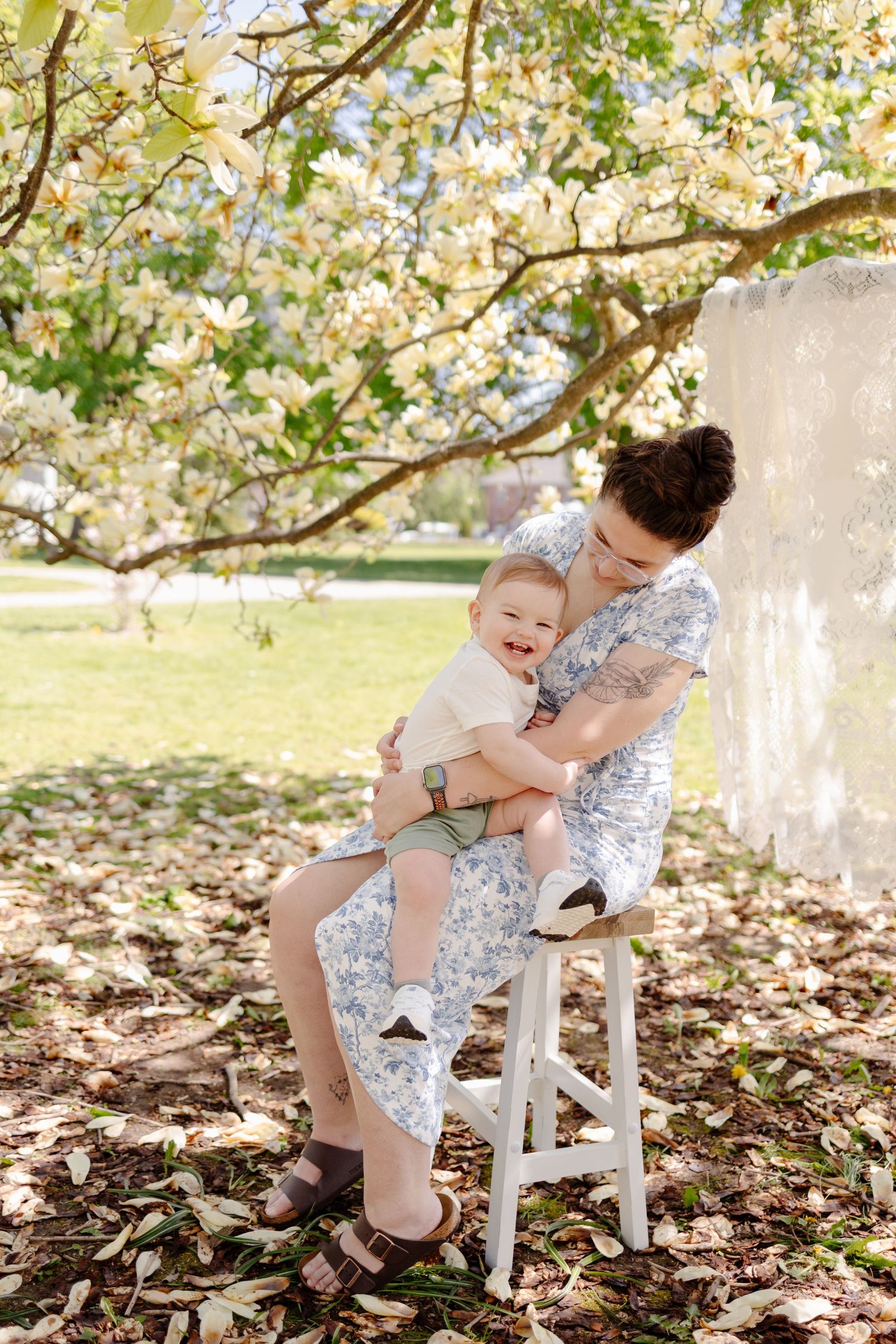 A tender moment shared under blooming spring magnolia trees with a white wooden chair on fallen leaves.