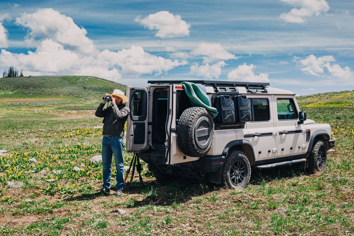 Photographer using a tripod camera beside a white off-road 4x4 vehicle in a green meadow under a cloudy sky.