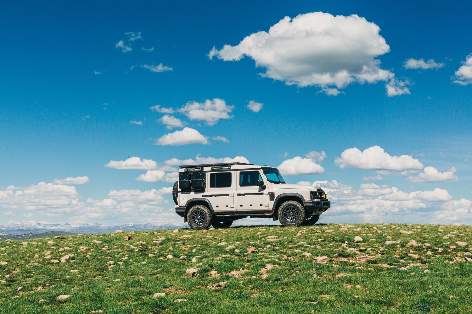 White off-road SUV with roof rack parked on green hilltop under blue sky with fluffy white clouds.