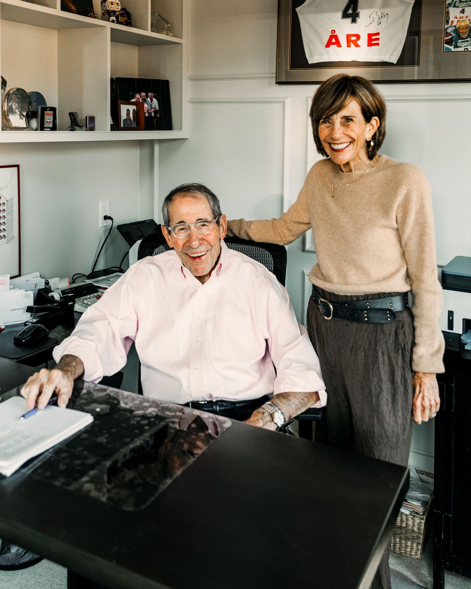 Smiling elderly couple posing together in a home office with bookshelves and framed memorabilia on the wall.