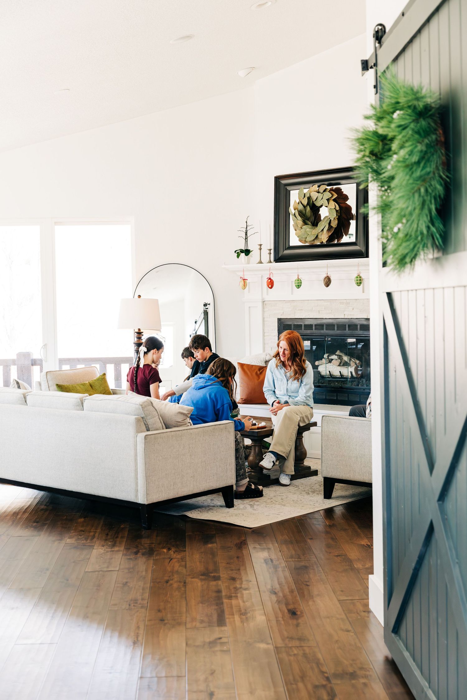 Family gathered in a cozy living room with fireplace, white sofa, hardwood floors, and Christmas wreaths decorating the space.