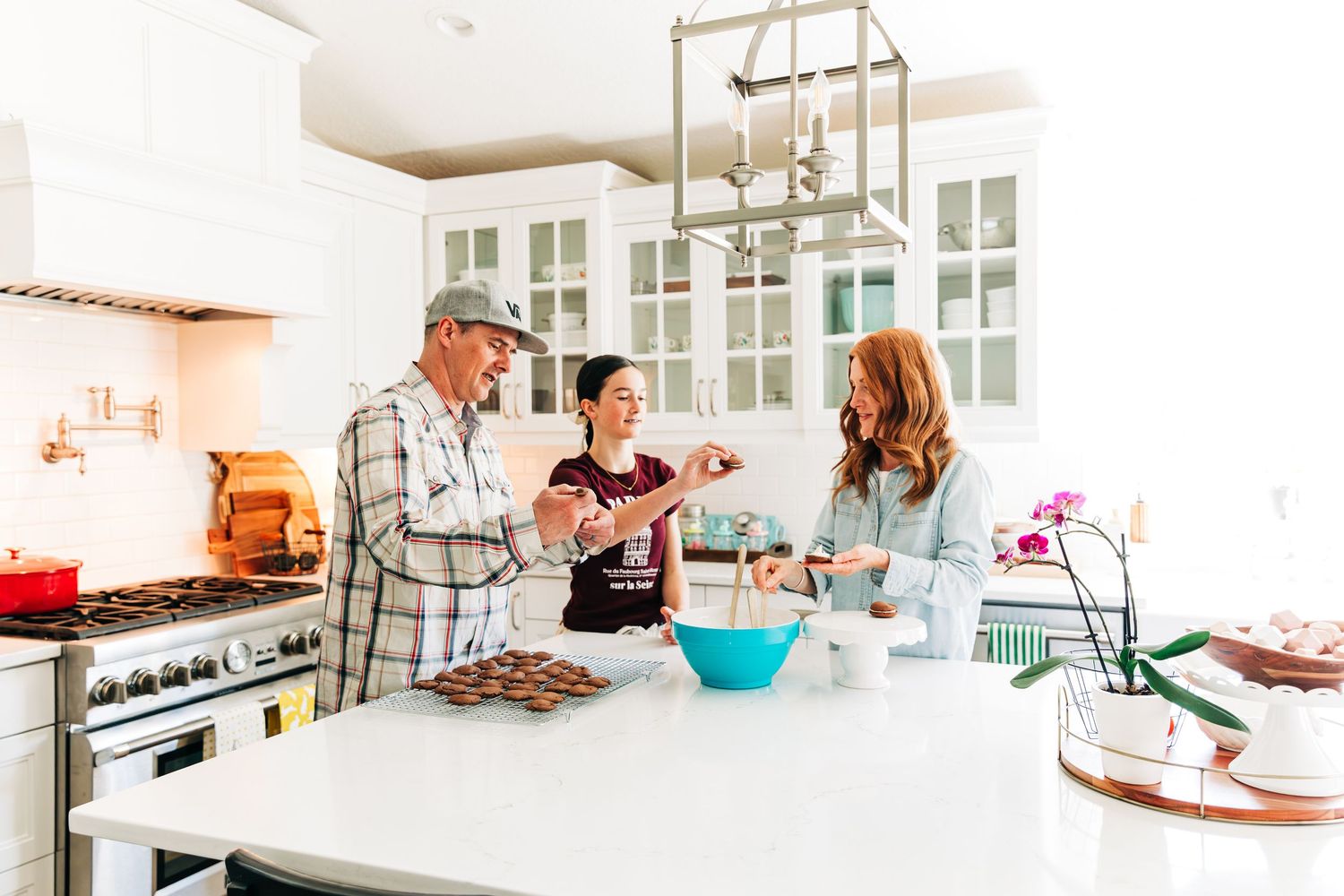Three people baking cookies together in a bright white modern kitchen with marble countertops and pendant lighting.