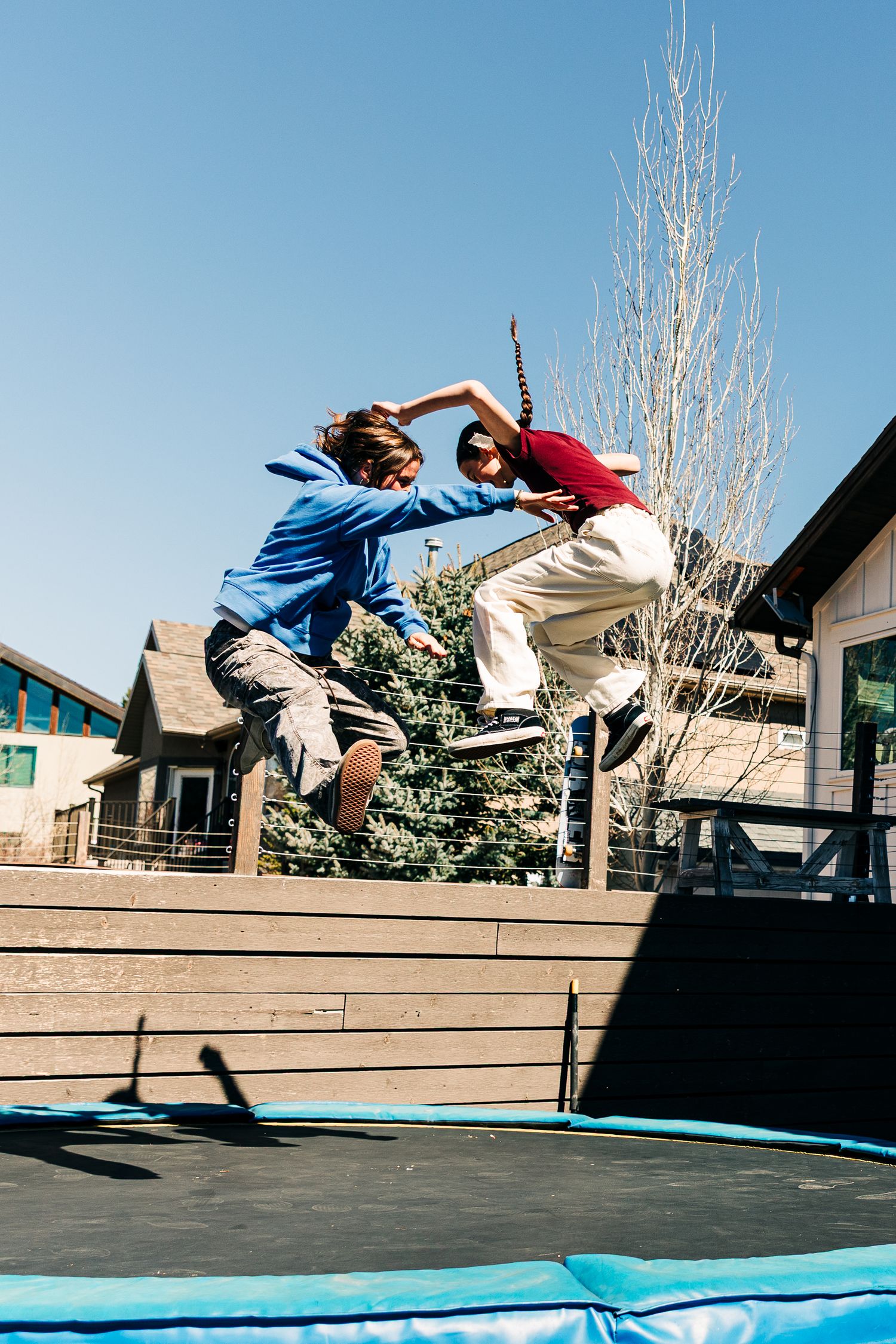 Two kids jumping on a backyard trampoline on a sunny day with houses and trees in the background.