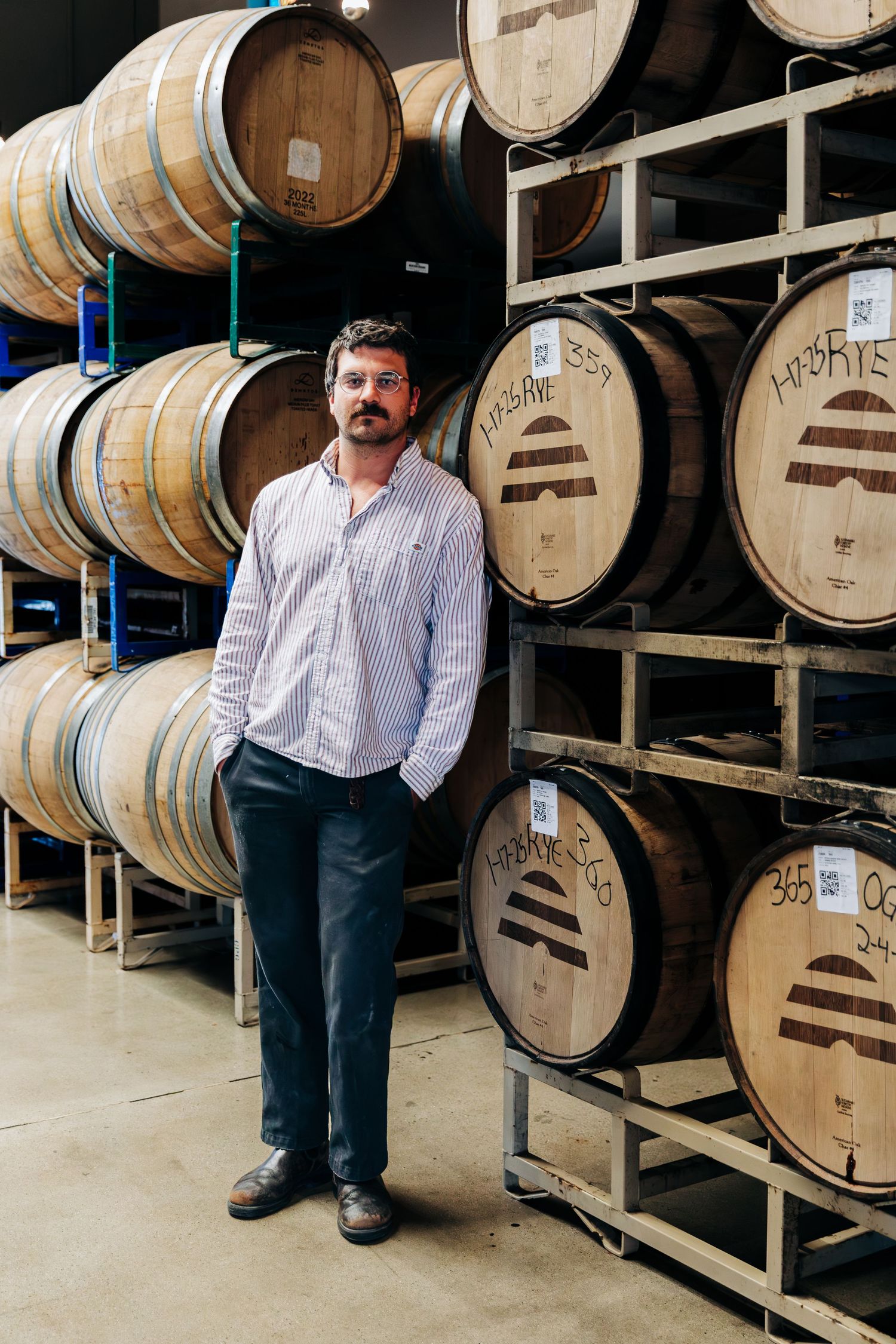 Man standing beside oak barrels in a distillery barrel aging room, wearing a striped shirt and dark pants.
