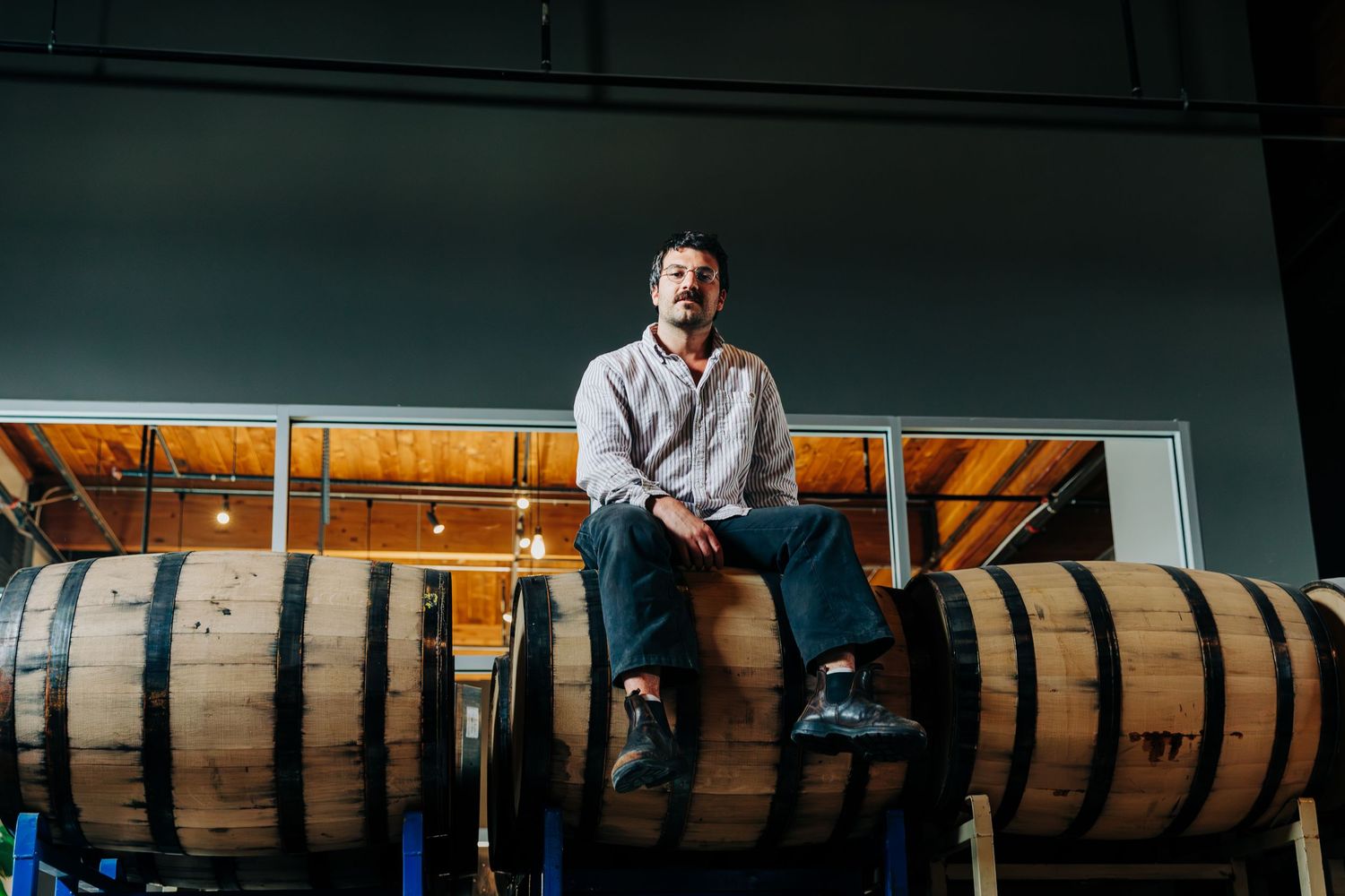 Man sitting on wooden barrels in a brewery or distillery warehouse, wearing casual attire.