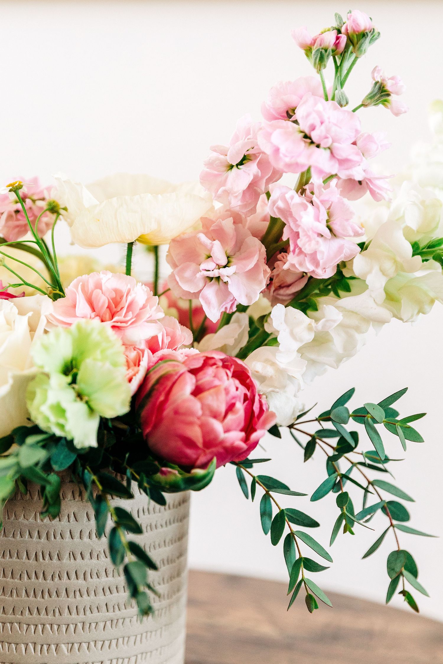 Elegant floral arrangement with pink peonies, roses, and eucalyptus in a white woven vase on a wooden surface.