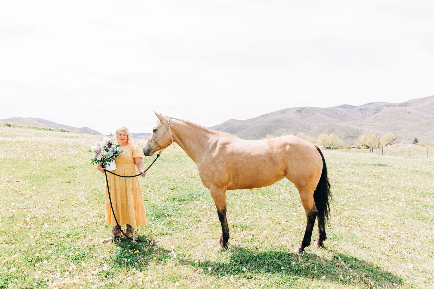 Woman in golden dress holds bouquet beside a tan horse in a green meadow with rolling hills backdrop.