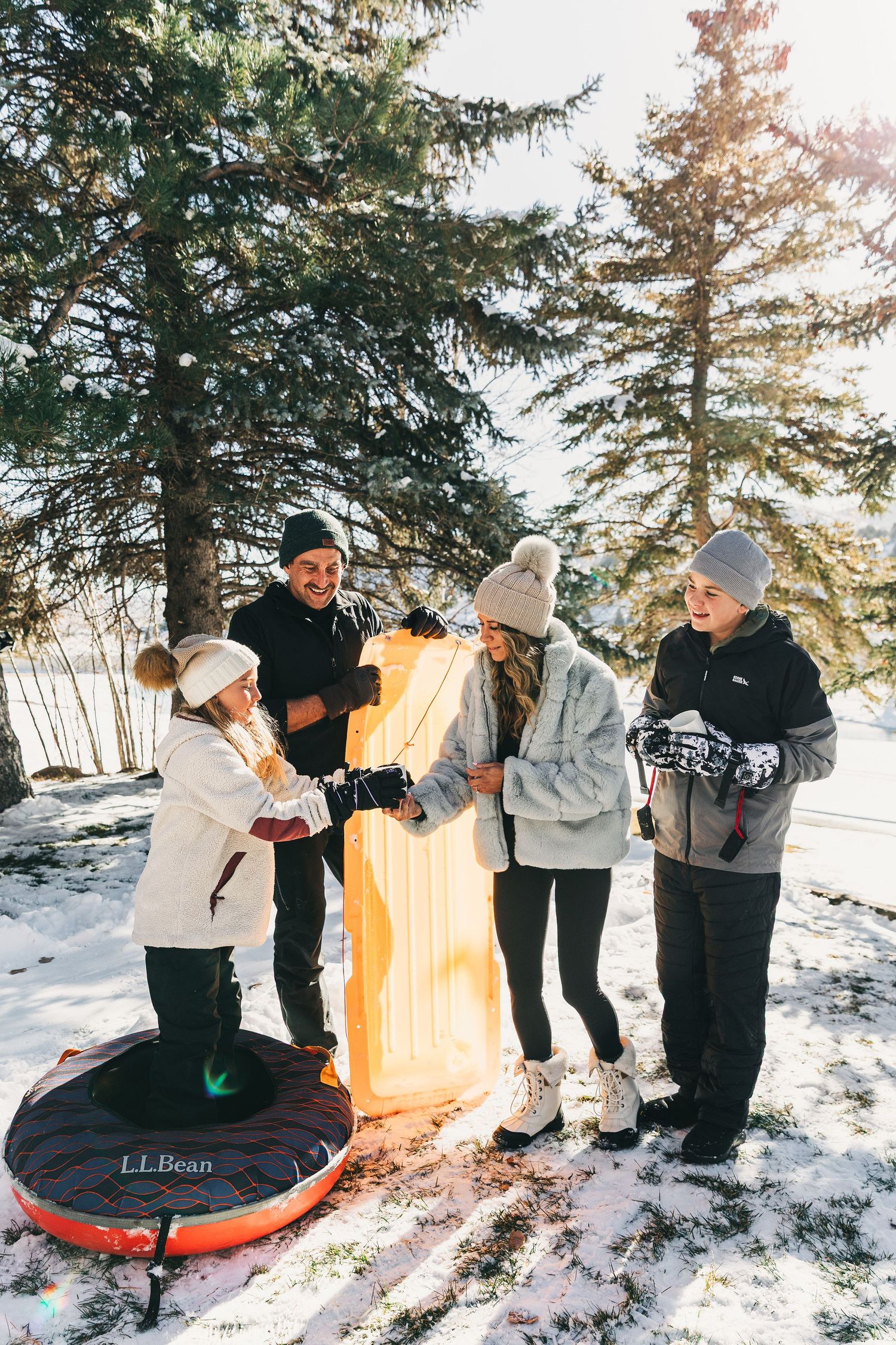 Family of four enjoying winter sledding with L.L.Bean sleds and snow tubes among snow-covered pine trees.