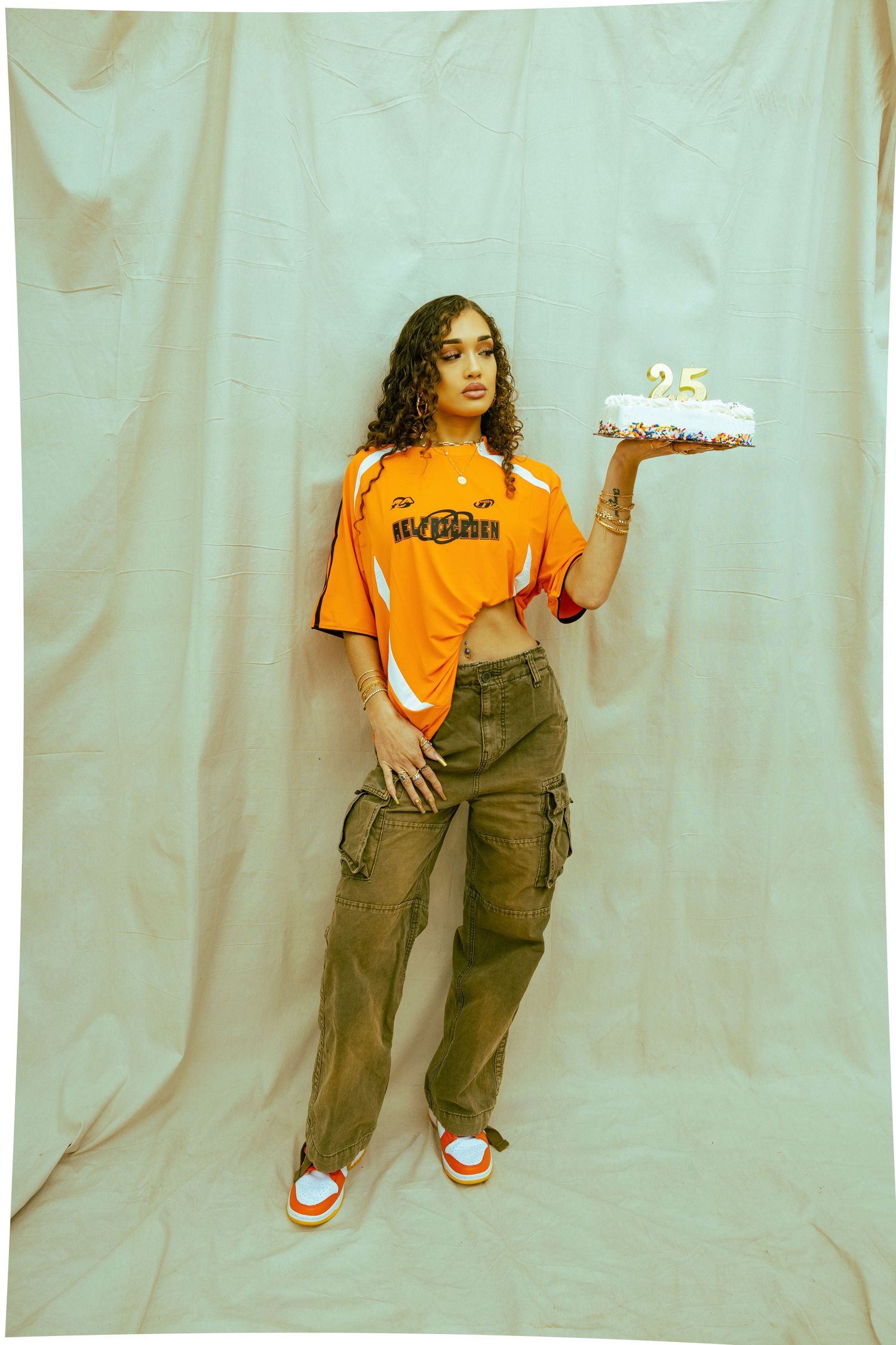 Person in orange shirt and cargo pants holds birthday cake against mint backdrop.